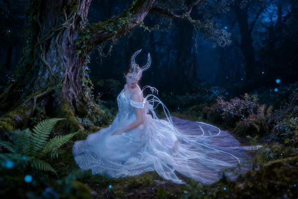 A horned, masked figure in an ethereal white costume sits beside a large tree in a moonlit forest, surrounded by blue light and glowing specks.