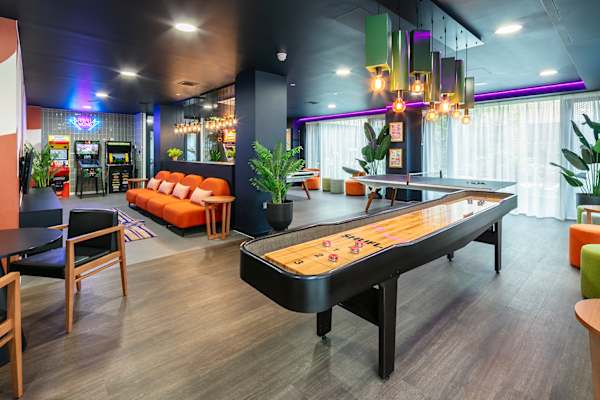 Games room at Archwood House with a shuffleboard table in the foreground and a table tennis table beyond. Pinball and arcade machines sit beneath a neon sign at the back, with an orange sofa, green and orange pouffes, plants and cluster pendant lights under a dark ceiling edged with purple lighting.