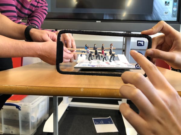 Image of student workshop where one student is holding a model phone pretending to take a photo, whilst other students use sticks with photos of people on which looks like they are walking across the screen. 