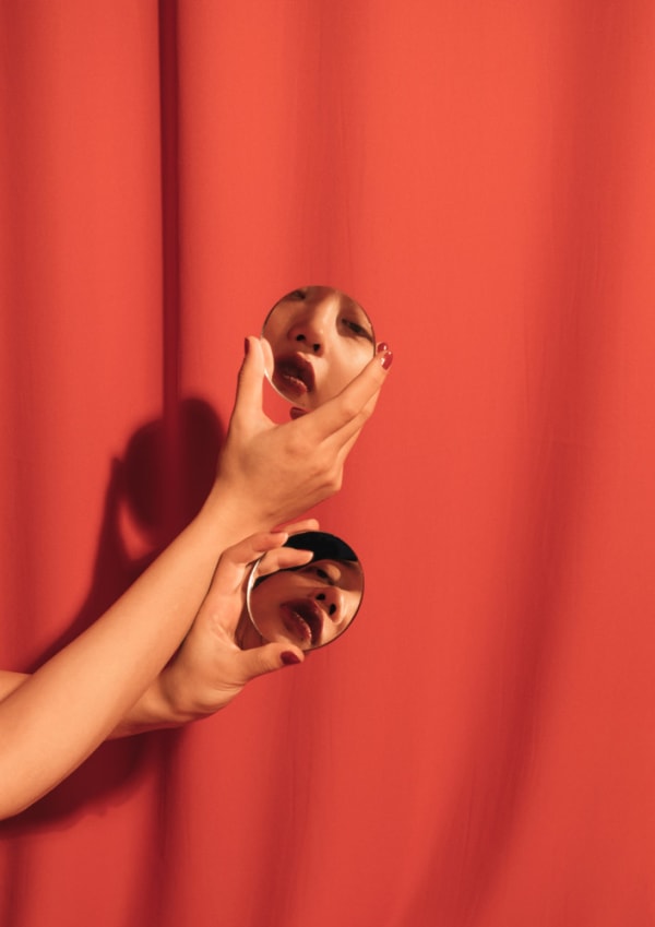Two compact mirrors are held by a pair of hands in front of a red curtain. The reflections depict different angles of a woman wearing red lipstick.