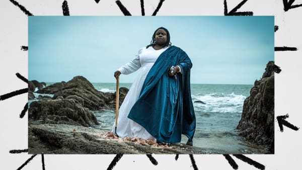 A person standing on the sand infront of the sea. They are wearing a floor length dress and cape and holding a tool 