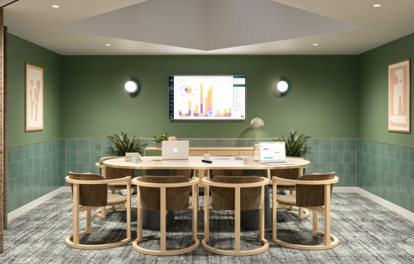 View of office showing a large round desk with a couple of laptops on it and  chairs around it, there is a wall mounted screen above the desk and the room is decorated with with dark green tiles, plants, and framed artwork.