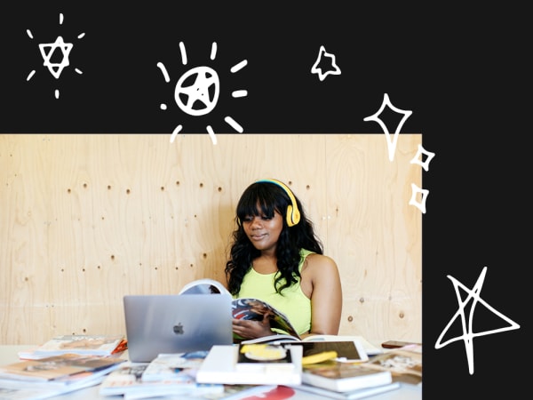 person reading a magazine, sitting in front of a desk with a laptop and several books spread out