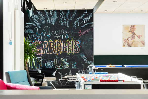 Shot of the common room, with bright neon student mural on chalkboard wall reading 'Welcome to Gardens house', foosball table a pool table, bunting and different seating areas.