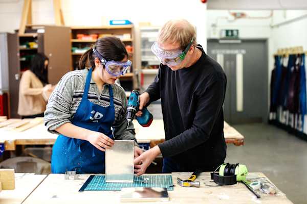 Two people wearing safety goggles drill a metal item in a workshop.