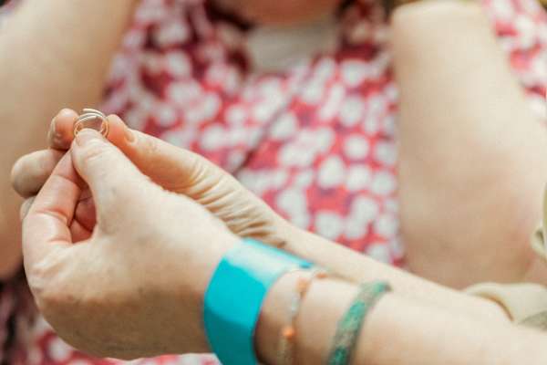 Close-up of hands holding a small coiled metal ring or jewellery component, with a blurred background showing a patterned red-and-white garment. The person is wearing a turquoise wristband and multiple bracelets on one wrist.