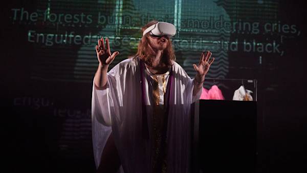Man dressed as a priest with his hands up wearing an AR headset with a digital screen behind him.