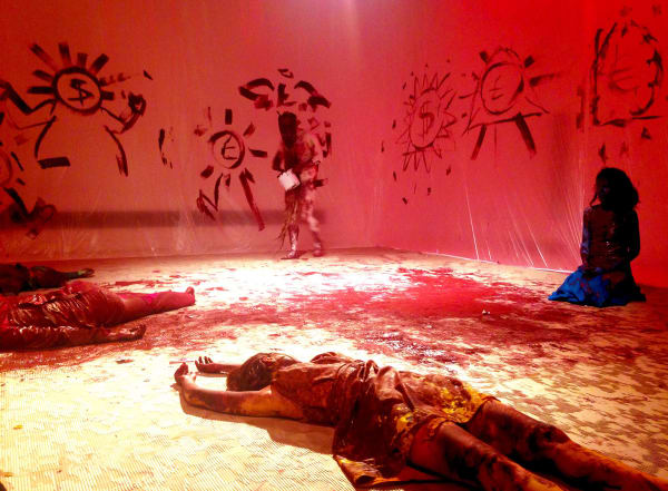 Photo of a room with red light, three people lying on the floor covered in red paint with one standing painting designs on the wall