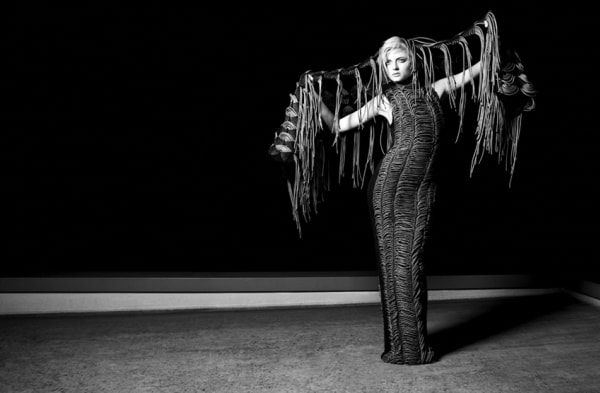 dramatic black and white photo of model posed arms outstretched in front of a black backdrop. Model wears a form fitting maxi tube dress, and a woven fringe garment behind hair and stretched over arms.