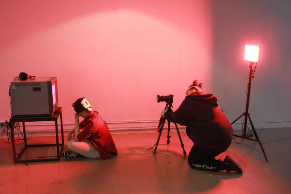 In a red-lit room a student films another student with a mask in front of an old TV