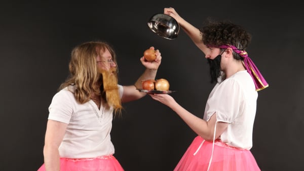 2 men with fake beards dressed in pink tutus and white tops holding a metal tray with onions on it.