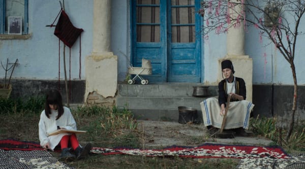 A richly detailed people scene from a film. Two characters are seated outside of a blue door, wearing striking garments.
