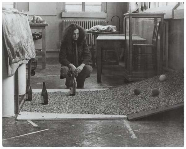 Black and white of a man crouching with an experimental assemblage in front of him, of two boards, covered in gravel, with three balls and three bottles