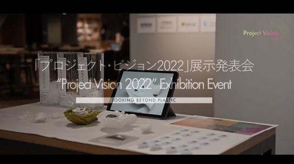 A title slide from an exhibition with the text: Project Vision 2022 Exhibition Event in English and Japanese. In the backgound is a table and poster board with innovative materials.