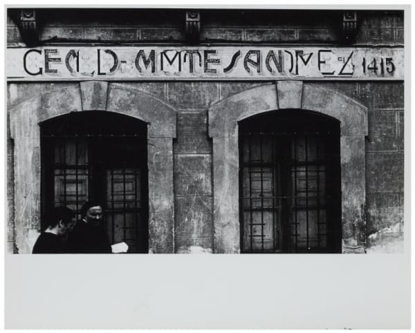 Black and white photograph of a shop sign where the lettering is peeling, showing different layers of lettering underneath. Two passers by are caught in the shot, walking in front of the building.