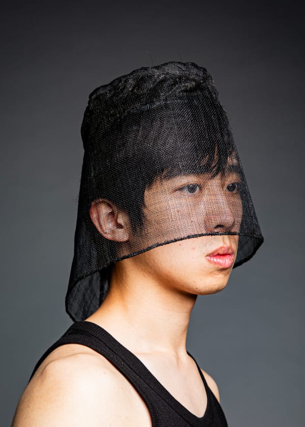 A model is shot from the shoulders up, they are wearing a black vest and a sheer black semi-opaque headpiece which comes down over the eyes and nose and is longer at the back, extending to the base of the neck