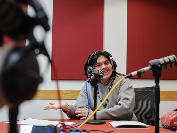 A student in a sound studio with headphones on smiling infront of a mic. They are in conversation with someone off camera. There is another person sitting across from them also with headphones