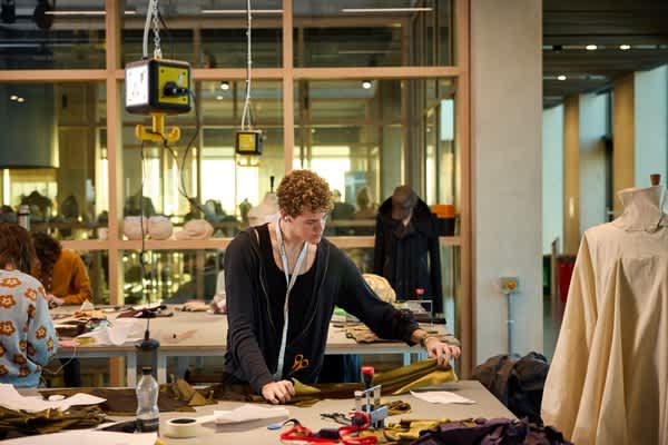 Student cutting and arranging fabric in a fashion design workspace at London College of Fashion, surrounded by garments, tools and pattern-making materials.