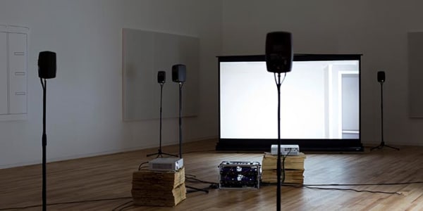 A gallery space with four speakers and a projection screen set up 