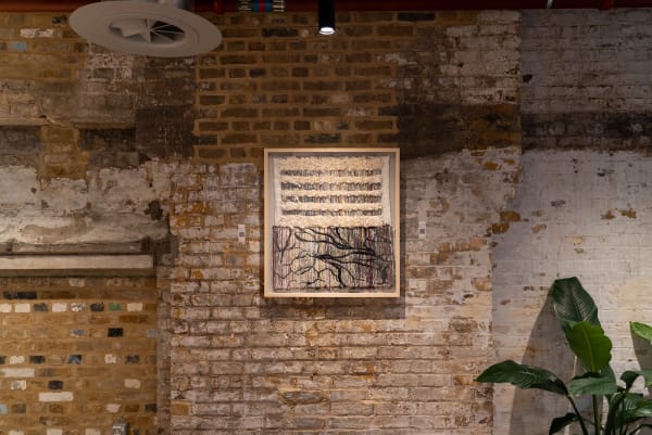 A square artwork piece framed and hanging on a brick wall. The artwork features woven textires and branch-like black lines