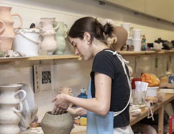 A student is standing at a working desk, shaping a vase. They are surrounded by examples of in progress ceramics work.