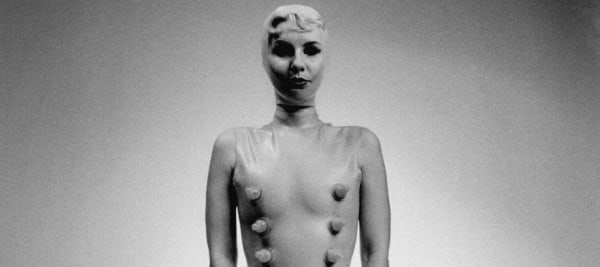 A grainy grey editorial shot of a female figure in a tight catsuit with hood and retro makeup 