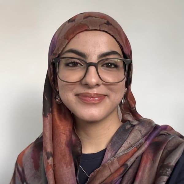 A student smiles at the camera in a head-and-shoulders portrait, wearing glasses and a patterned headscarf against a plain background.