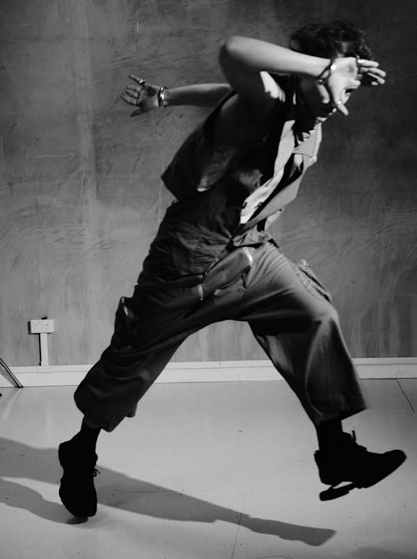 Black-and-white fashion photograph of a person in a dynamic mid-stride pose, wearing a sleeveless vest and wide-leg trousers in a studio setting.