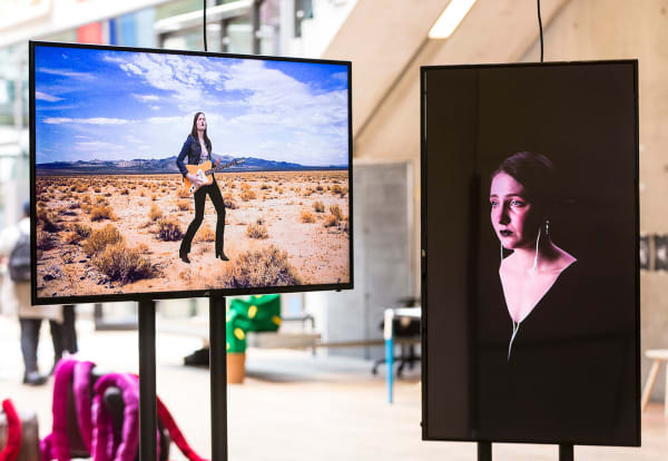 1 portrait and one landscape screen set on frames in an open space during an exhibition. The screens are showing different scenes of video with people as the centre of the screen. The background is bright and naturally lit, you can see a staircase and people walking around.