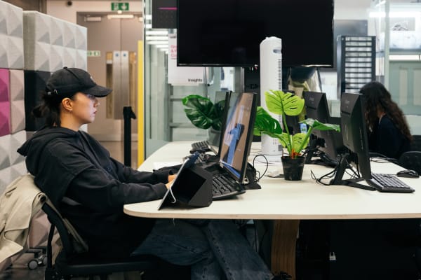 Shot from the side, a student in a baseball cap is using a keyboard and widescreen computer in a booth with a plant.