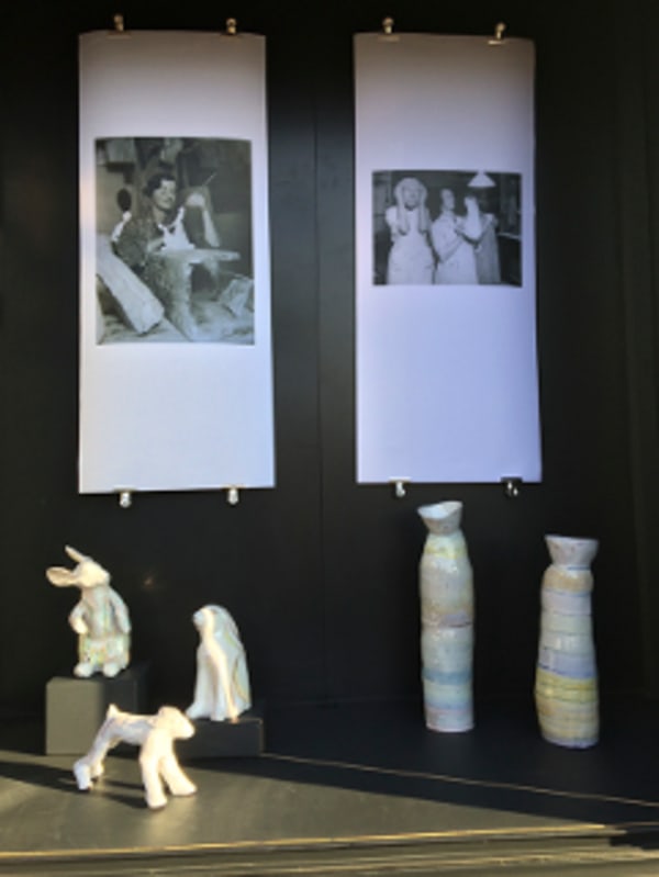 Ceramic figures in the shape of playful animals and vessels on display, framed by 2 hanging black and white photographic prints.