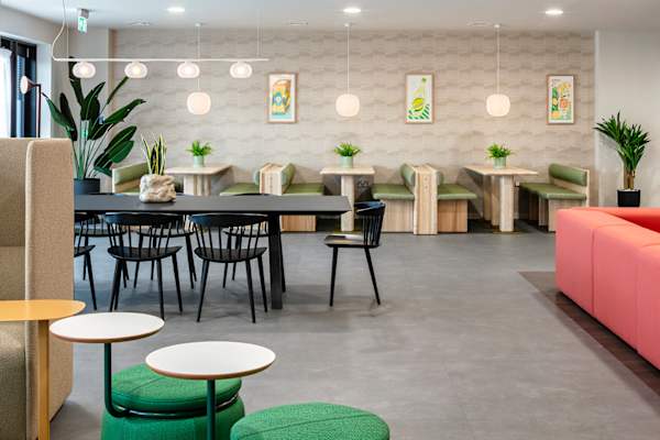 Bright social space at Archwood House with green upholstered booth seating and pale wood tables against a patterned wall hung with three colourful drinks-themed prints. In front, a long black dining table with black chairs, green pouffes with small round side tables, and part of a coral sofa.
