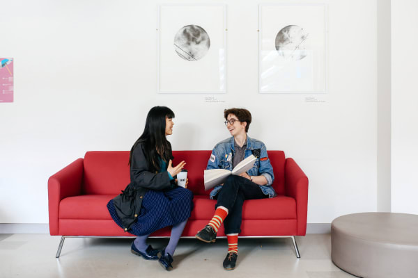 Two students having a conversation, sat on a red sofa. 