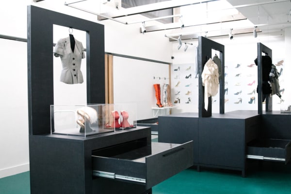 exhibition space with black display cabinets, showcasing hanging womenswear blouses, and perspex boxes with ornaments, in the distance a few stiletto knee high boots. on the far wall a collage print of footwear designs.