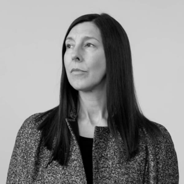 Professor Jane Harris, Director of UAL Fashion, Textiles and Technology Institute is standing in front of a white background, looking off into the distance to the left, she has long dark hair and is wearing a wool coat