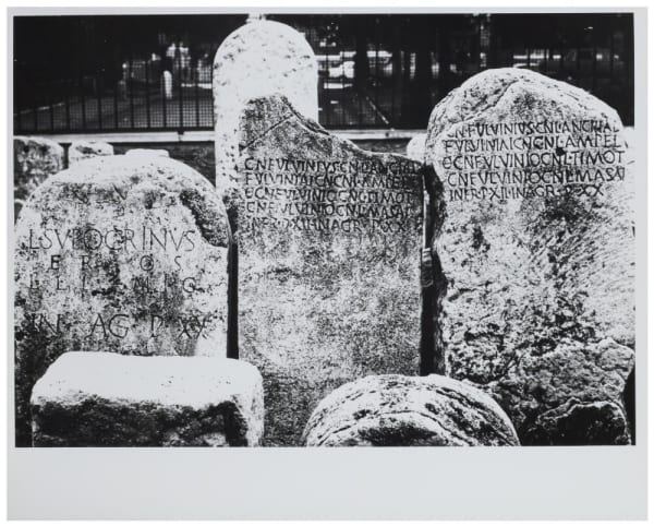 Black and white photograph of rounded stones that might be tombstones, with lettering in Latin carved into them. They are broken and look quite old