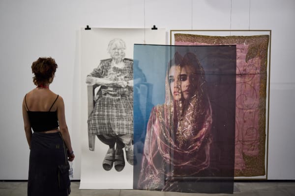 An observer is standing looking at a trio of large-scale hanging photographic, drawing and textile artworks, the photo is a stylised shot of a woman in a veil, next to a pink embroidered sari cloth with gold trims. Behind this is a detailed drawing of a seated woman with crossed legs wearing tartan. 