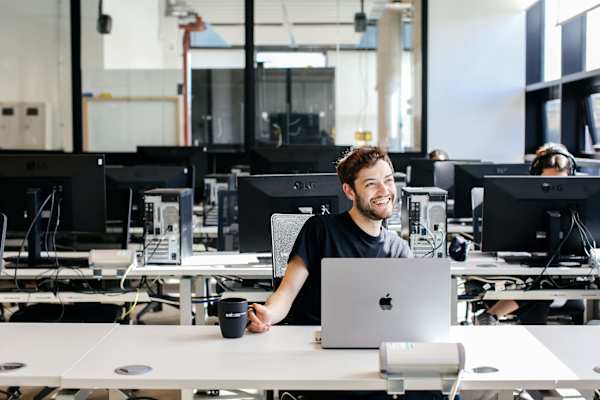 A person sitting in front of a laptop smiles while holding a mug.