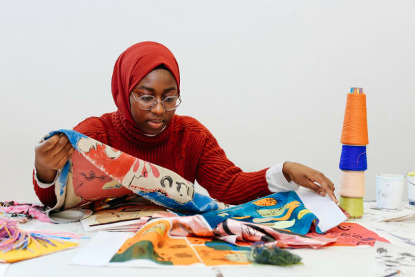 Black female student sitting at workbench looking at various brightly coloured fabrics