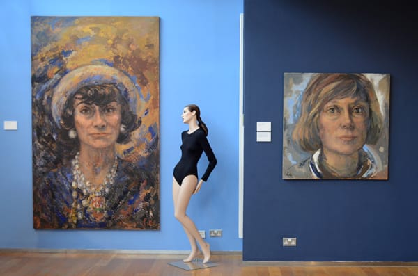 exhibition view of two multicoloured large portrait paintings of Coco Chanel against two blue walls. In between both paintings a mannequin posed wearing a navy unitard.