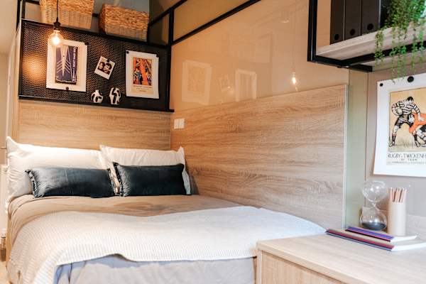 Close-up of a neatly made bed with white bedding and dark velvet cushions, set against a wooden headboard panel with overhead shelving displaying framed prints, wicker baskets, and decorative objects.