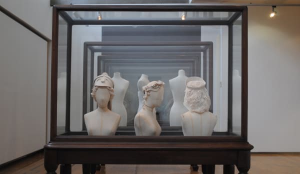 Installation view of three fabric mannequin head and shoulders, wearing different prototype millinery. Left to right, mannequin facing towards camera in a officer style hat, middle mannequin facing right wearing an elegant headdress, right mannequin facing away from camera wearing a hairnet.