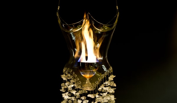 close up of a glass sculpted dress with a flame inside. The top half shaped like a corset, and the bottom half made of wired glass elements hanging down.