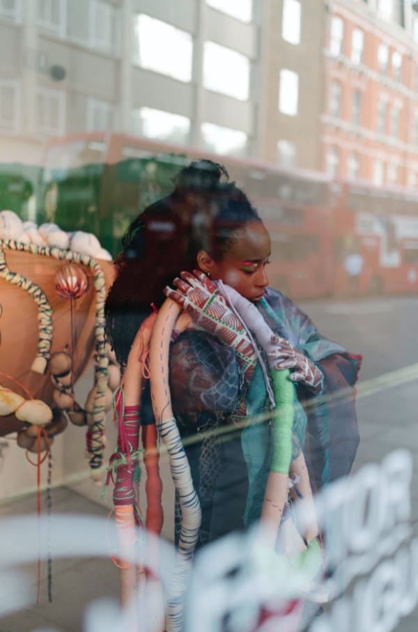 photo of Alethia through a store window, posed with ropes covered in multicoloured thread, and textured animal print gloves.