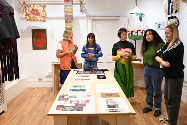 Image shows 5 people standing round a table with textile-based artwork in the background and on the table. The people are engaged in discussion as they admire the work. 