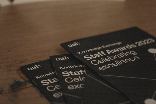 Wooden table with 4 black cover booklets piled on top of each other, the booklets have 'UAL Knowledge Exchnage Staff awards 2023, celebrating excellence' on the cover