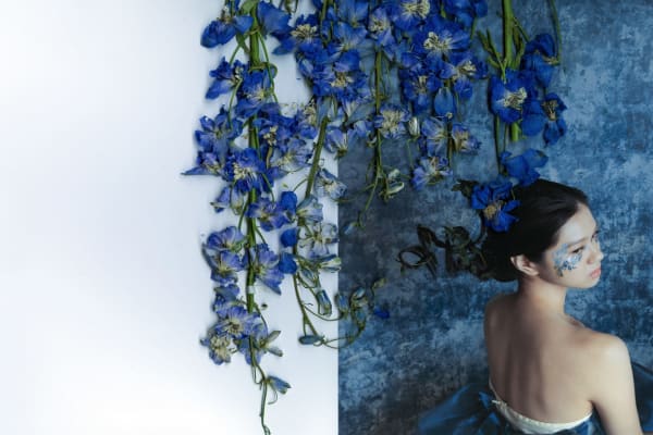 Photo of woman with floral arrangement through hair, that goes onto the wall. Colours are purple and blue tones, with flowers matching wall colours and woman's make up.