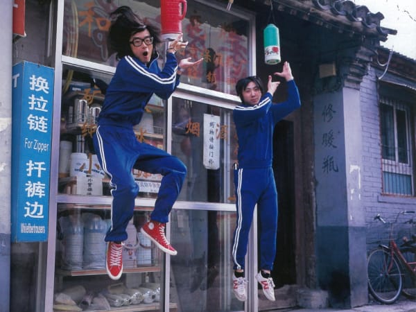 Two men in blue striped tracksuits jumping in the air in front of an Asian shop.