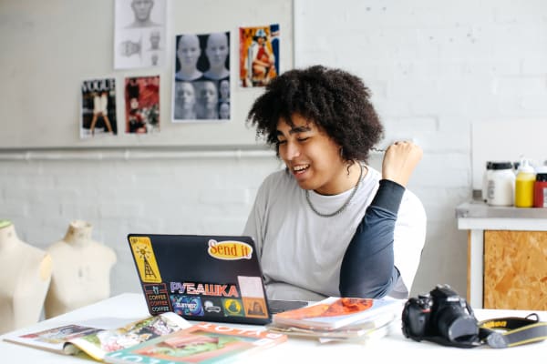 Issac is wearing a white t-shirt over a dark long sleeved shirt. They are sitting at a table in a fashion studio looking at their laptop. They are twirling a strand of their curly hair in between their fingers.