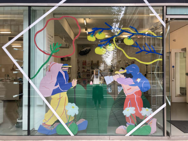Street-facing view of a window display at 'not just a shop' featuring a vibrant illustration applied to the glass. The artwork shows two colourful, cartoon-style figures: one on the left wearing a backpack and hiking gear, and another on the right holding binoculars and kneeling to observe. Large abstract flowers and fruit-laden branches extend around them, framing the scene. The figures appear to be exploring or birdwatching, evoking a sense of adventure and nature. Inside the shop, shelves and displays are visible, along with a reflection of the opposite street buildings in the glass.